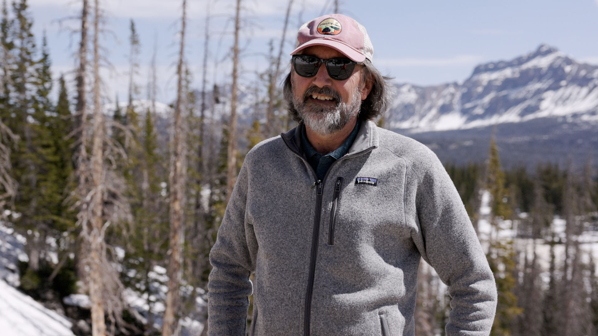 Man in a cap and gray fleece sweater stands in front of snowy mountain landscape