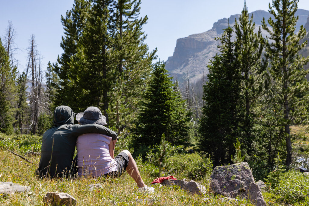 Two hikers are seen from behind, sitting in a meadow, with one wrapping an arm around the other