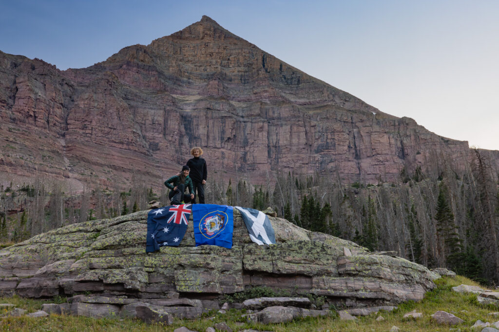 Two women stand on a rock outcropping with flags, below a towering triangular mountain peak